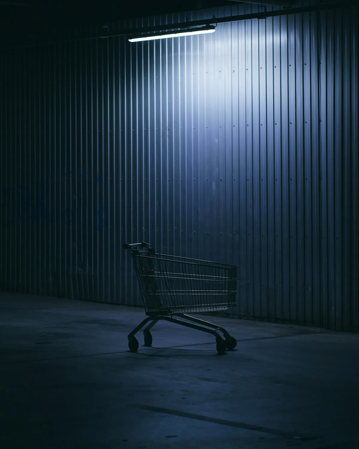 Empty shopping cart in a dark room, lit from above against a ribbed metal wall